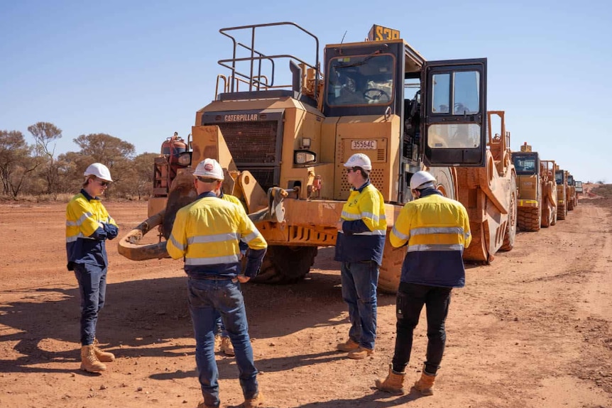 Four people in high-vis construction gear in front of heavy machinery.