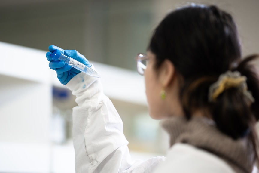 A laboratory assistant in a white lab coat holding a transparent vessel against a white blurry background.