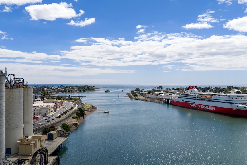 An aerial view of a calm ocean port, with industrial silos on the left bank and a large passenger ferry berthed at right.