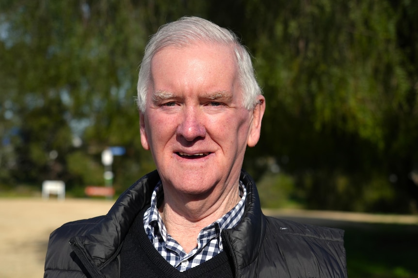 A man with grey hair wearing a blue chequered shirt and a black jacket stands in front of a green tree on a sunny day.