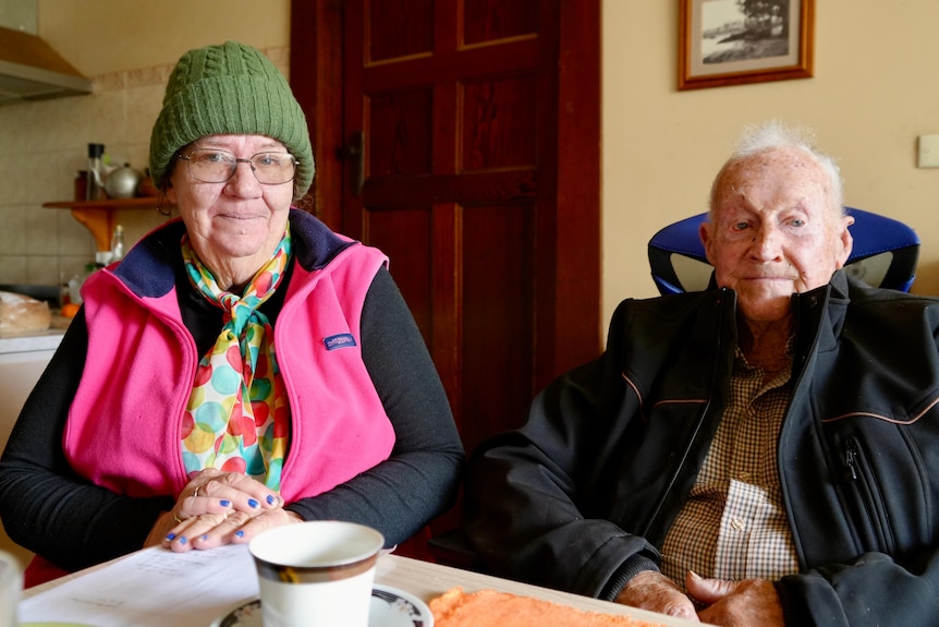 a lady in a pink vest and beanie with glasses and a older man with grey hair and a black jacket smiling at the camera 