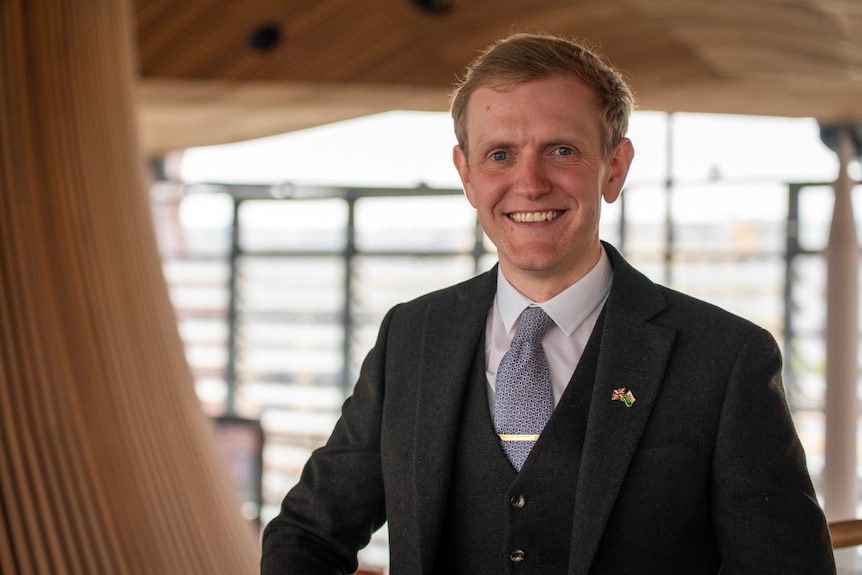 A man in a suit smiles at the camera while standing inside a large building.