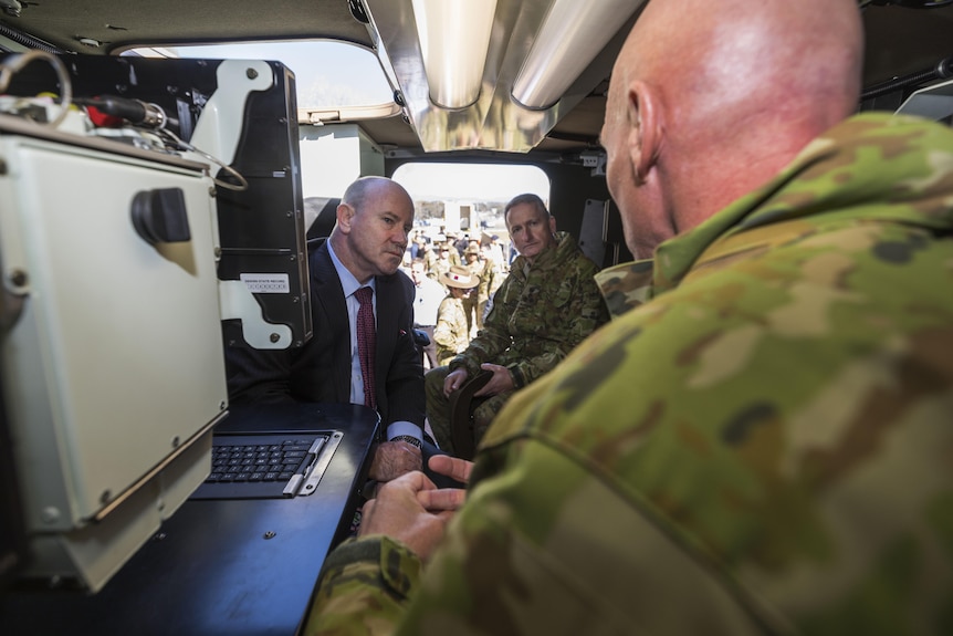 Three men sitting in a small space, one with his back to the camera in army uniform, another two looking at him while he speaks