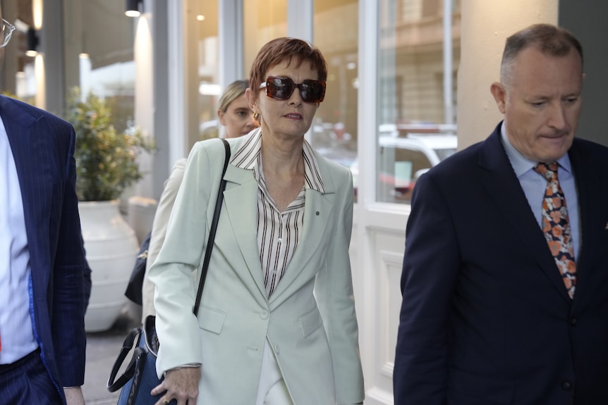 Gail wears a pale blazer and striped shirt and dark glasses. She walks alongside two men in suits.
