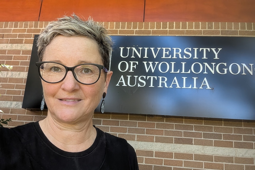 A white woman with short fair hair and black glasses standing in front of a University of Wollongong sign