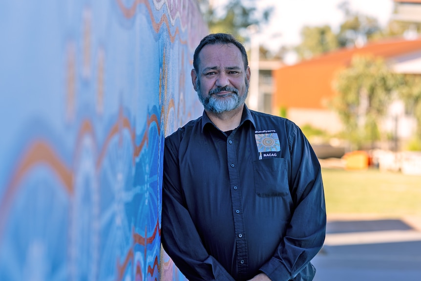 Jamie is wearing a black buttoned shirt leaning against a painted wall with Aboriginal art
