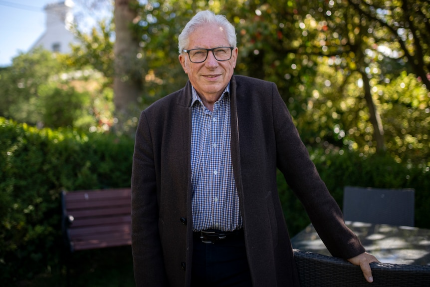 A gentleman in a suit and blue checkered shirt smiles with green hedges in the background.