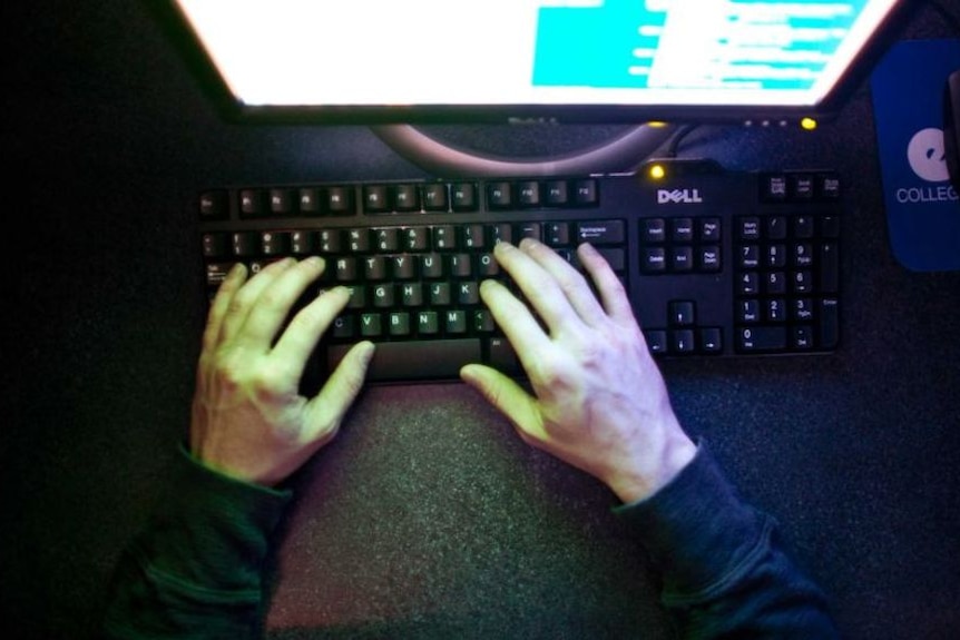 A person's hands are illuminated by the pale glow of a computer screen, as they type in a dark room.