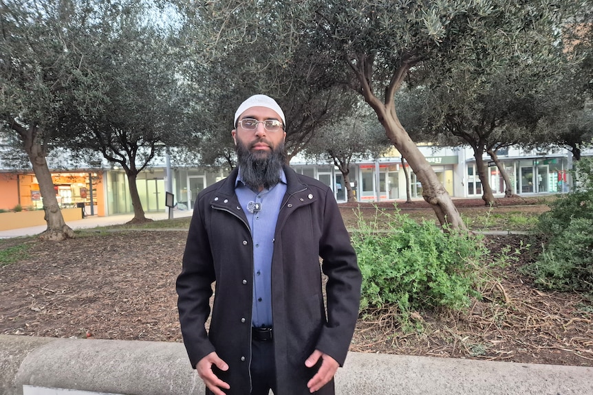 A man stands in front of trees with a neutral expression