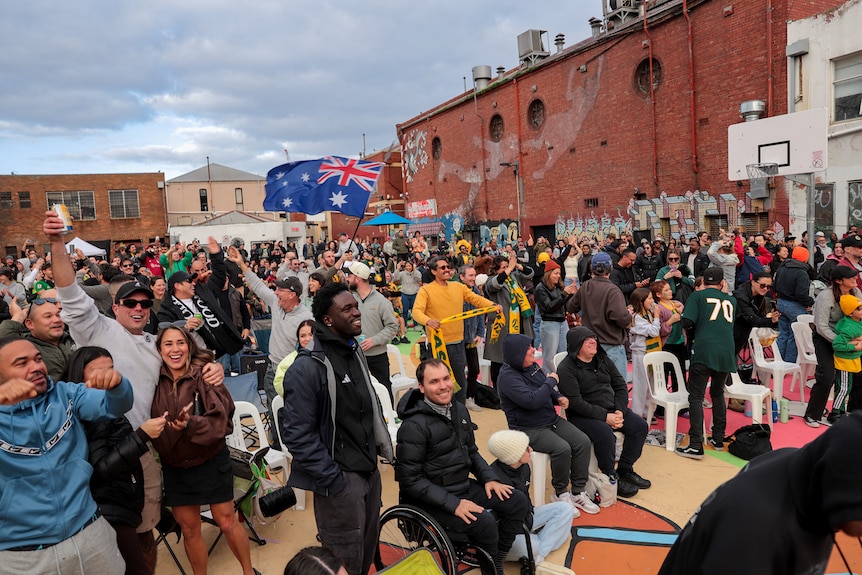 A group of people standing and sitting and looking ahead, some are wearing green and gold or holding Australian flag.