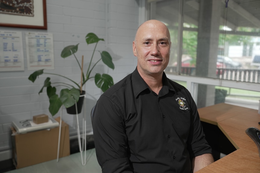 A man in a black collared shirt sits in an office