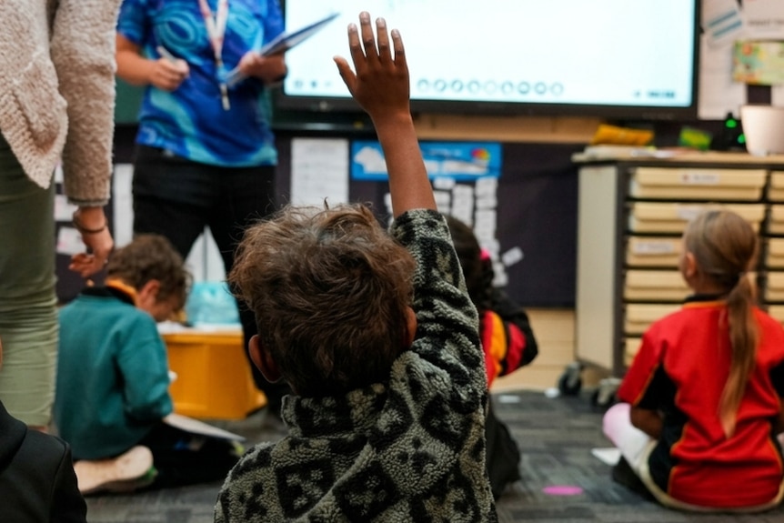Children sitting on the ground in a classroom in front of screen and teacher. A student has his hand in the air.