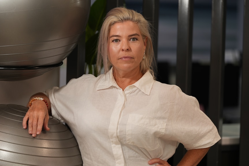A blonde woman standing alongside exercise equipment, she wears a serious facial expression while looking at the camera.