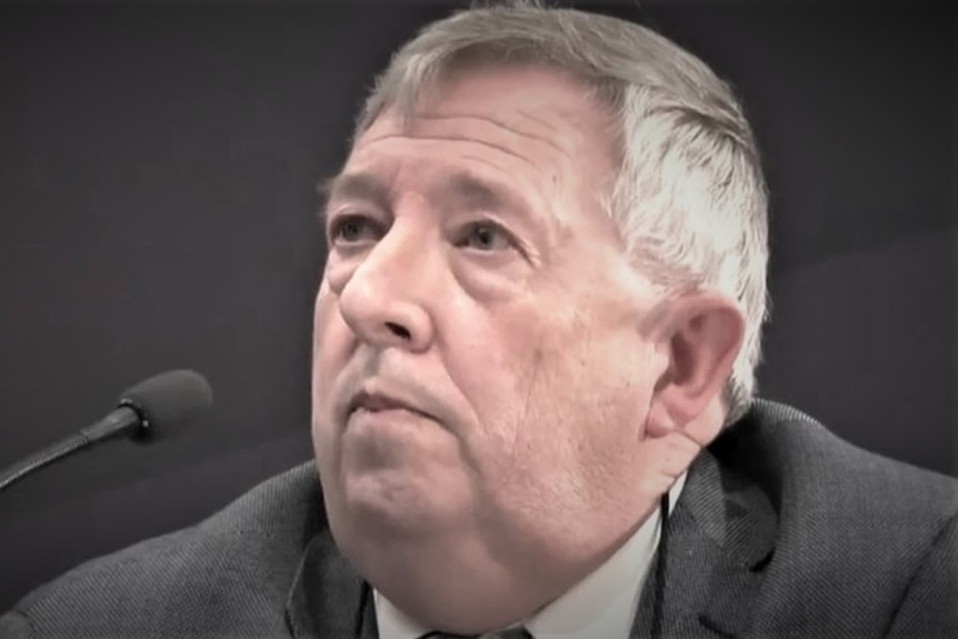A middle-ged man with grey hair wears a suit as he sits behind a microphone.