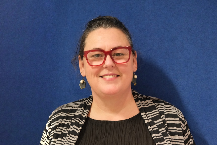 A woman wearing red glasses with her hair tied back standing in front of a blue backdrop