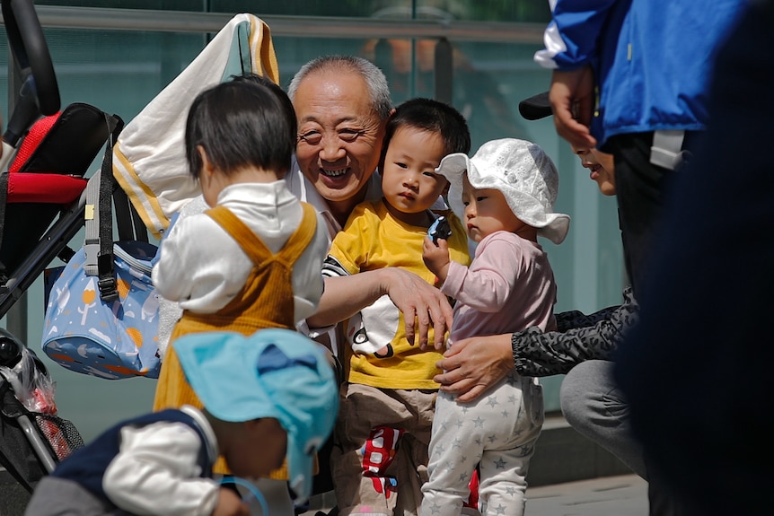 An elderly man smiles widely as he squats to hug and play with three small children in front of an office building.