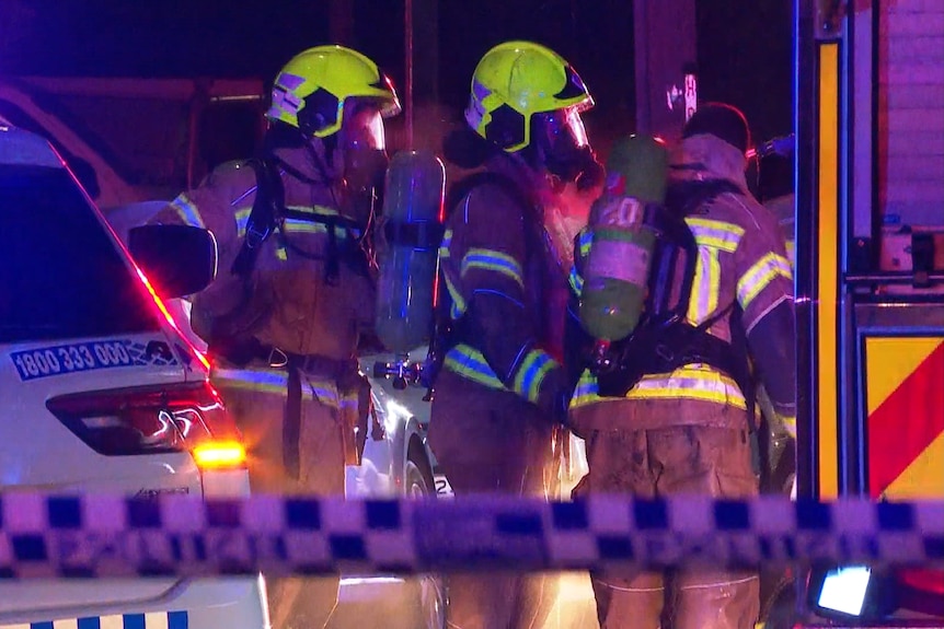 Three firefighters in protective gear wearing oxygen masks gather between a police car and fire truck.