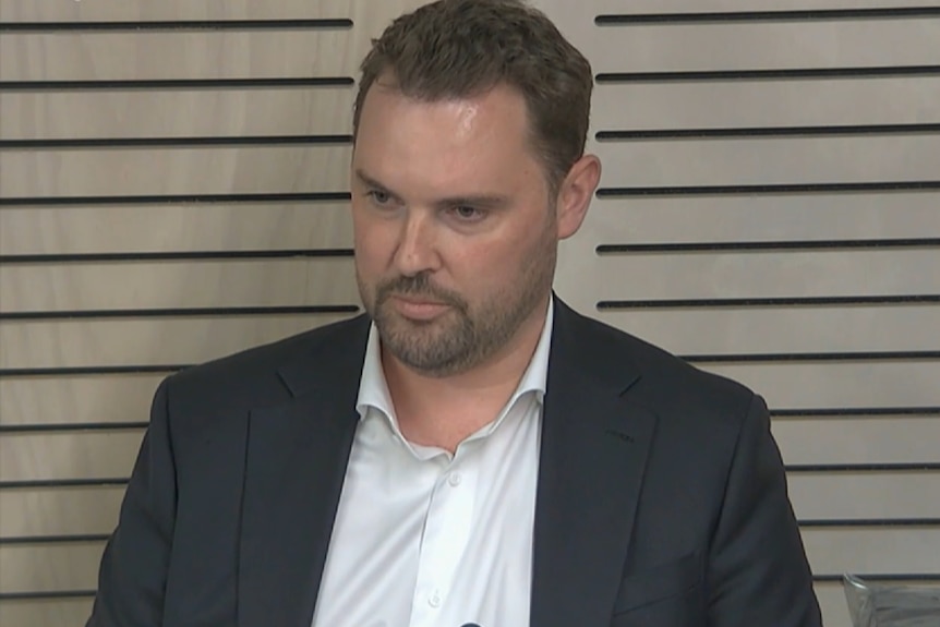 Mr Greenwood in a black blazer and white collared shirt sits at a desk with a serious expression.