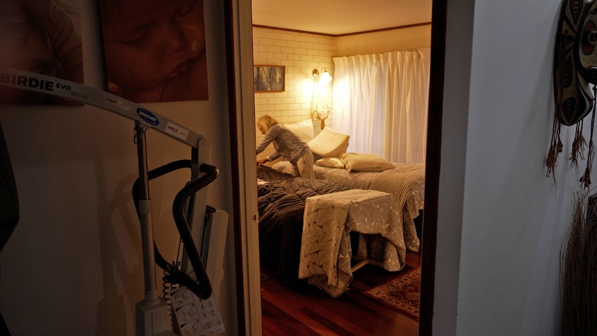 A photo shot from a hallway of a woman making a bed in a bedroom. Mobility equipment is in the hallway.