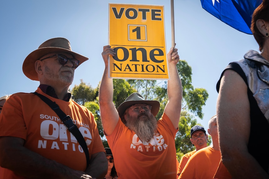 A man with a hat and beard holds a sign which reads Vote 1 One Nation