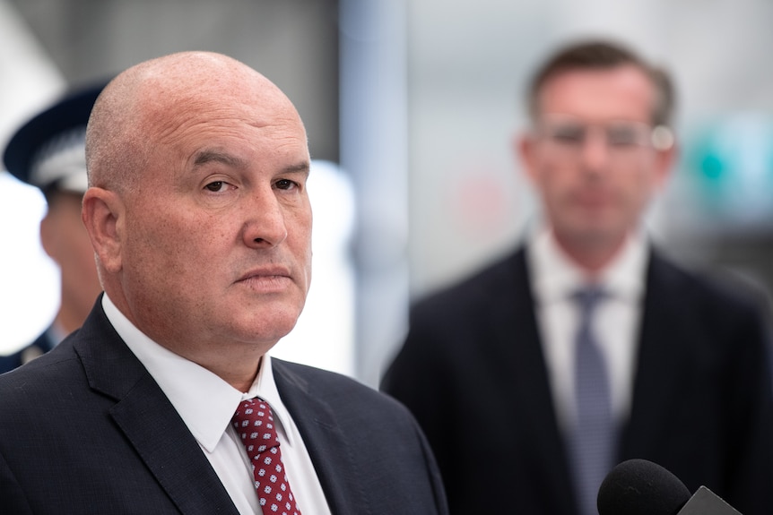 a man standing talking to the media with another man wearing glasses standing behind him