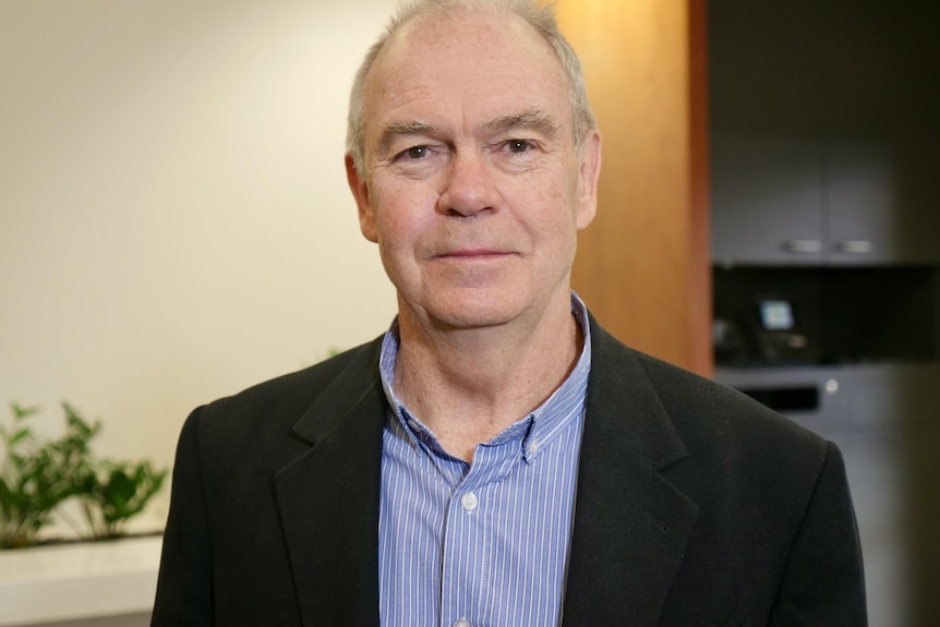 A headshot of an elderly man wearing a blue shirt and a dark jacket, with plants in the background