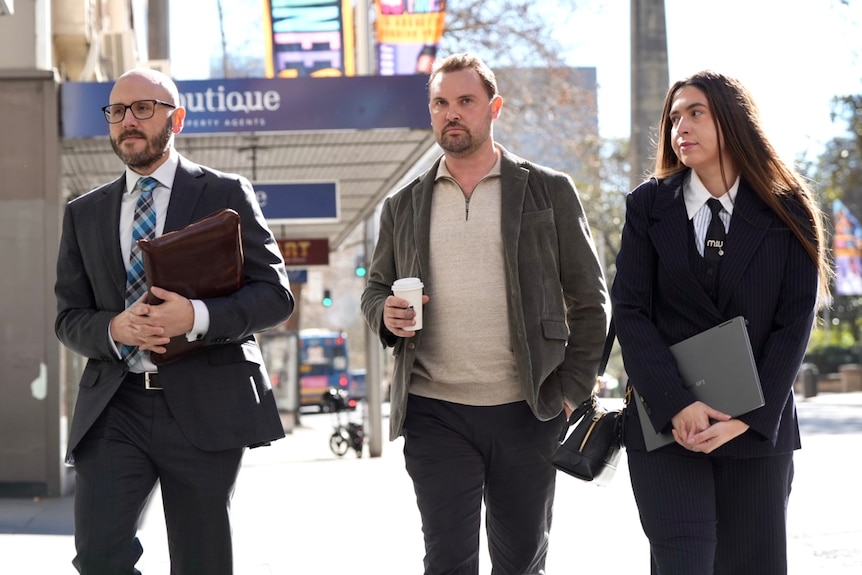 Mr Greenwood with facial hair wearing a brown jacket and white shirt holds a coffee while walking.