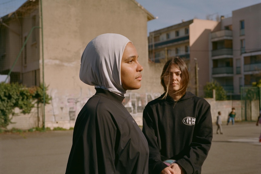 A photo of two women, one with a head scarf, stand near buildings in a paved area.