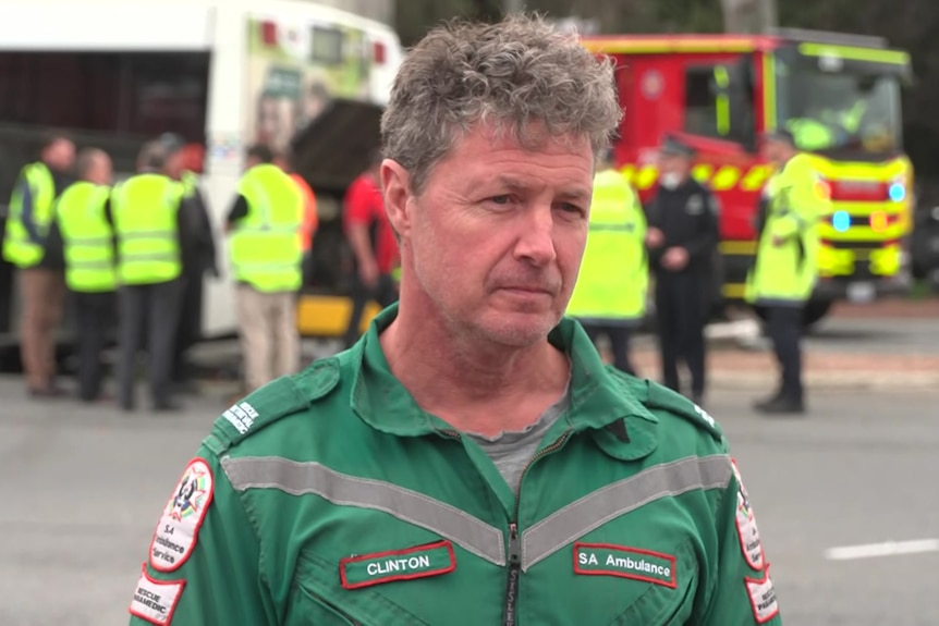 A man in a green SA Ambulance officer uniform .stands before a crash scene.