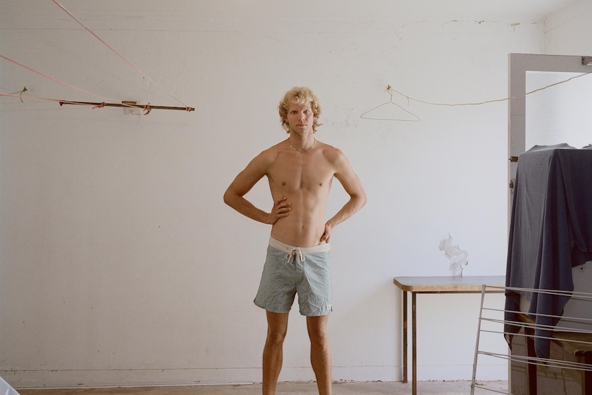 A photo of a young blonde man in shorts standing in a bare room with hands on hips.