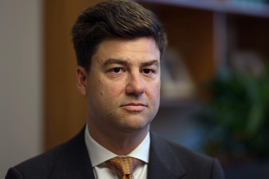 Sam Rae, dressed in suit and tie, sits in an office with a serious expression.