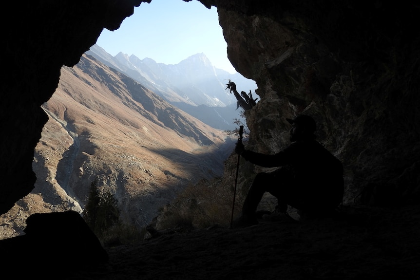 Dark outline of a person carrying a hiking stick sitting in a cave, watching a vista of tall sunny mountains.
