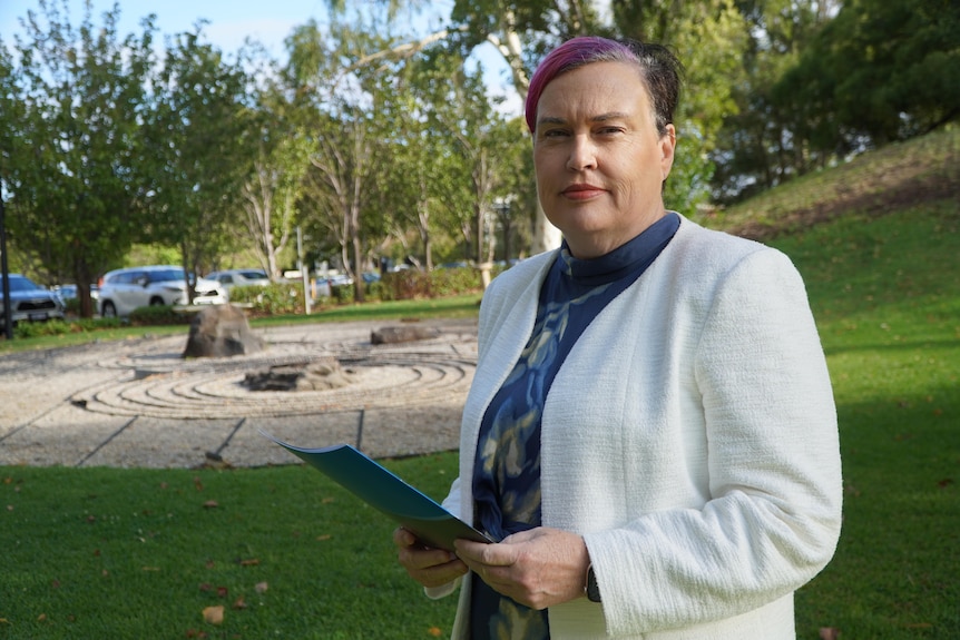 Trish Blake stands holding a booklet in a park.