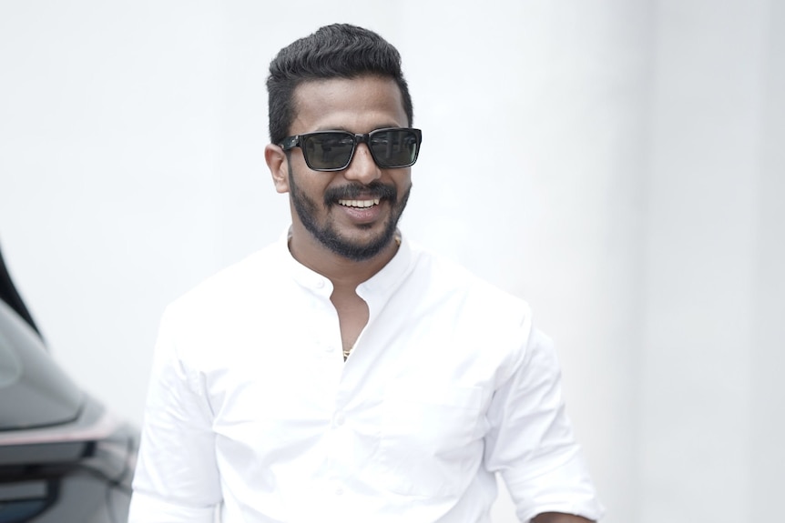 A man with a dark complexion smiling, wearing a white dress shirt and sunglasses.
