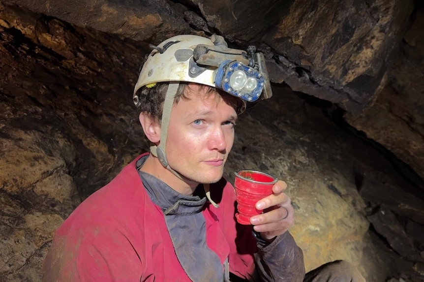 A man in a helmet sitting in a cave, holding a cup up to his mouth. 