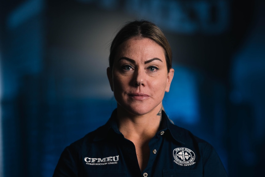 Woman with short hair tucked behind ears stares staunchly at the camera, hands held in front of her, in CFMEU uniform