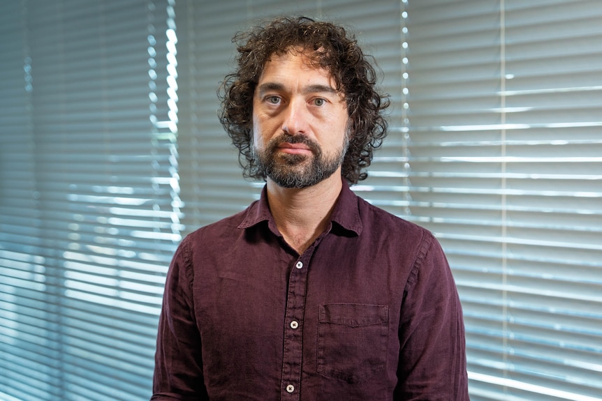 A man with black hair and a purple shirt looks at the camera