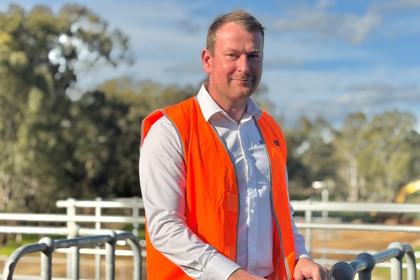 A man in a long sleeve white shirt and orange safety vest stands near metal fencing.