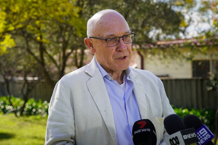 A man wearing glasses standing outdoors and speaking to the media