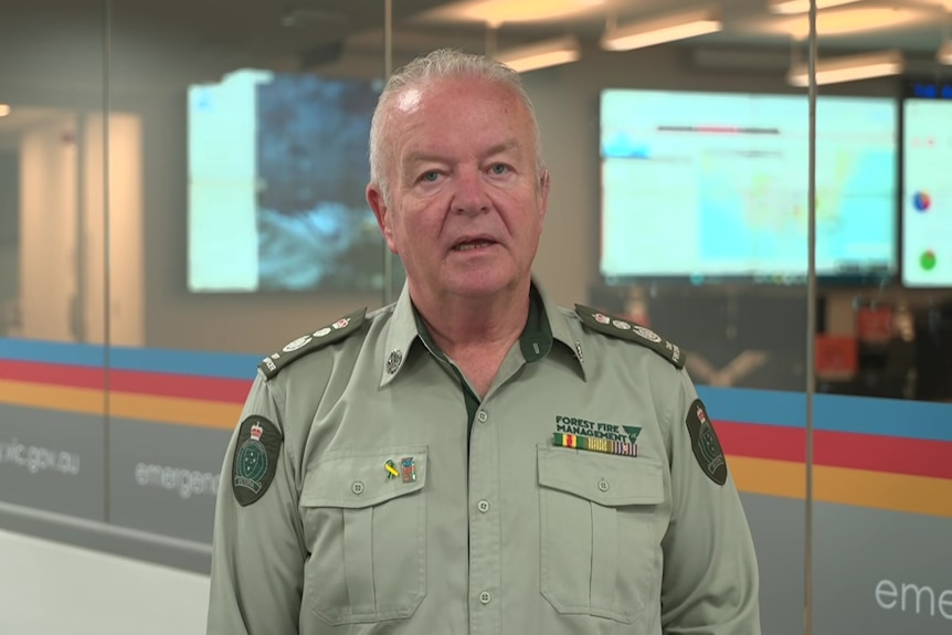 A man with grey hair in a light green collared shirt with badges stands near a glass wall and large computer screens.