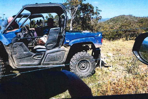 A man in a blue work shirt leaning on a blue farm buggy in bushland.