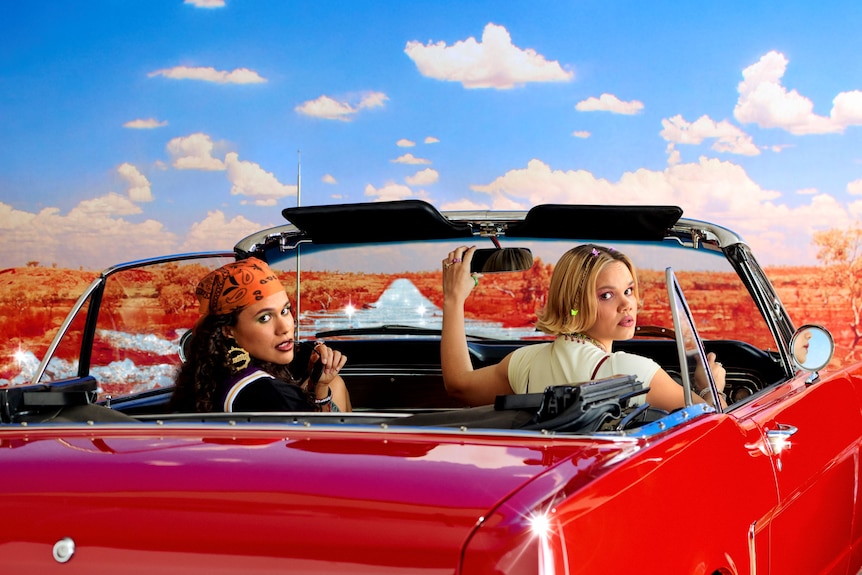 Ruby Henaway and Lila McGuire, two young Indigenous women, are sitting in a red convertible. They both look back at the camera.