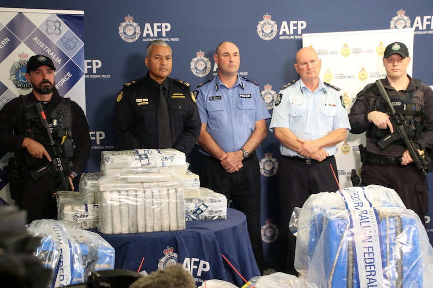 A line of police, some armed, standing behind plastic wrapped packaged piles of cocaine.
