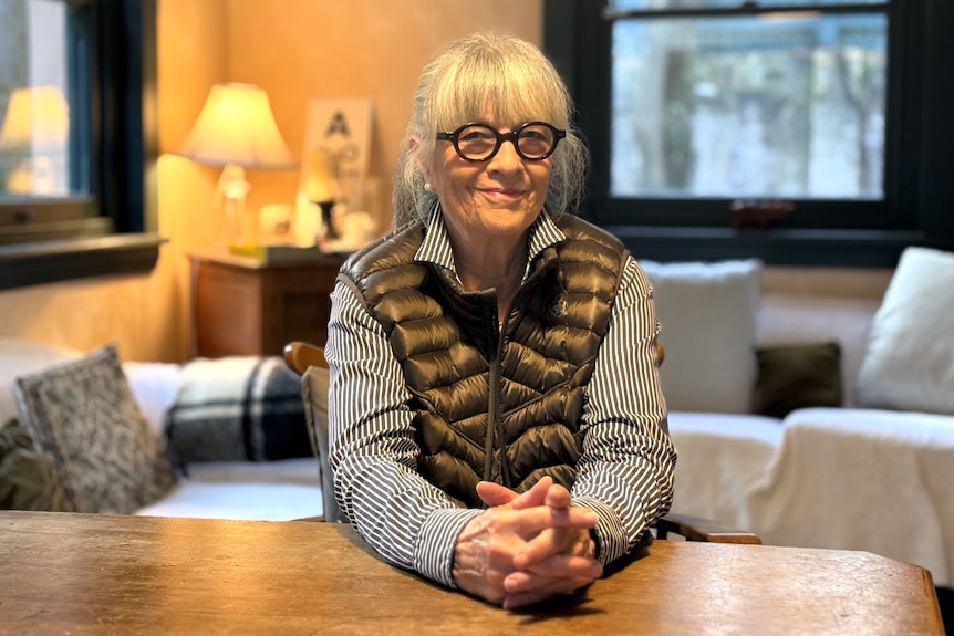 A woman with silver hair in a ponytail and dark-rimmed glasses smiles while sitting in a cosy living room. 