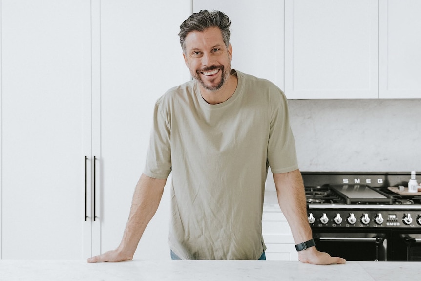 Man in beige t-shirt leaning on kitchen bench with wide smile