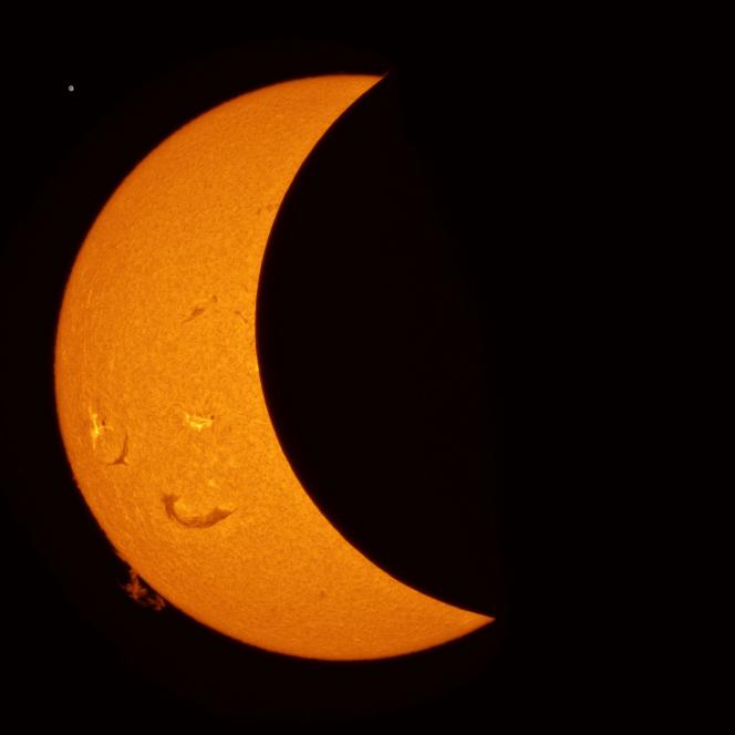 Les explications du photographe. Croissant de soleil à 19h55… 18 minute avant le maximum. L’observation de l’éclipse est faite en H-Alpha c’est à dire avec un filtrage ultra étroit centré sur la longueur d’onde 656,28 nm. On observe sur le limbe (périphérie solaire) une protubérance « à 8 h ». Il s’agit d’une immense structure de gaz ionisé (plasma) suspendue au-dessus de la surface du Soleil et guidée par ses champs magnétiques.Des filaments sont vu en projection sur le surface solaire, il s’agit des mêmes structures que les protubérances.Bien que plus chauds que la surface solaire, les filaments apparaissent noirs par effet d’absorption et de diffusion de la lumière H-alpha sous-jacente. La protubérance est claire, bien plus chaude que le vide spatial. On voit deux taches, phénomènes surtout photosphérique ( » la surface » proprement dite), elles sont entourées de fibrilles qui se disposent sous l’influence des champs magnétiques. Notez également des zones plus brillantes et chaudes dites plages faculaires, et des spicules qui donnent au disque un aspect de buissonnant