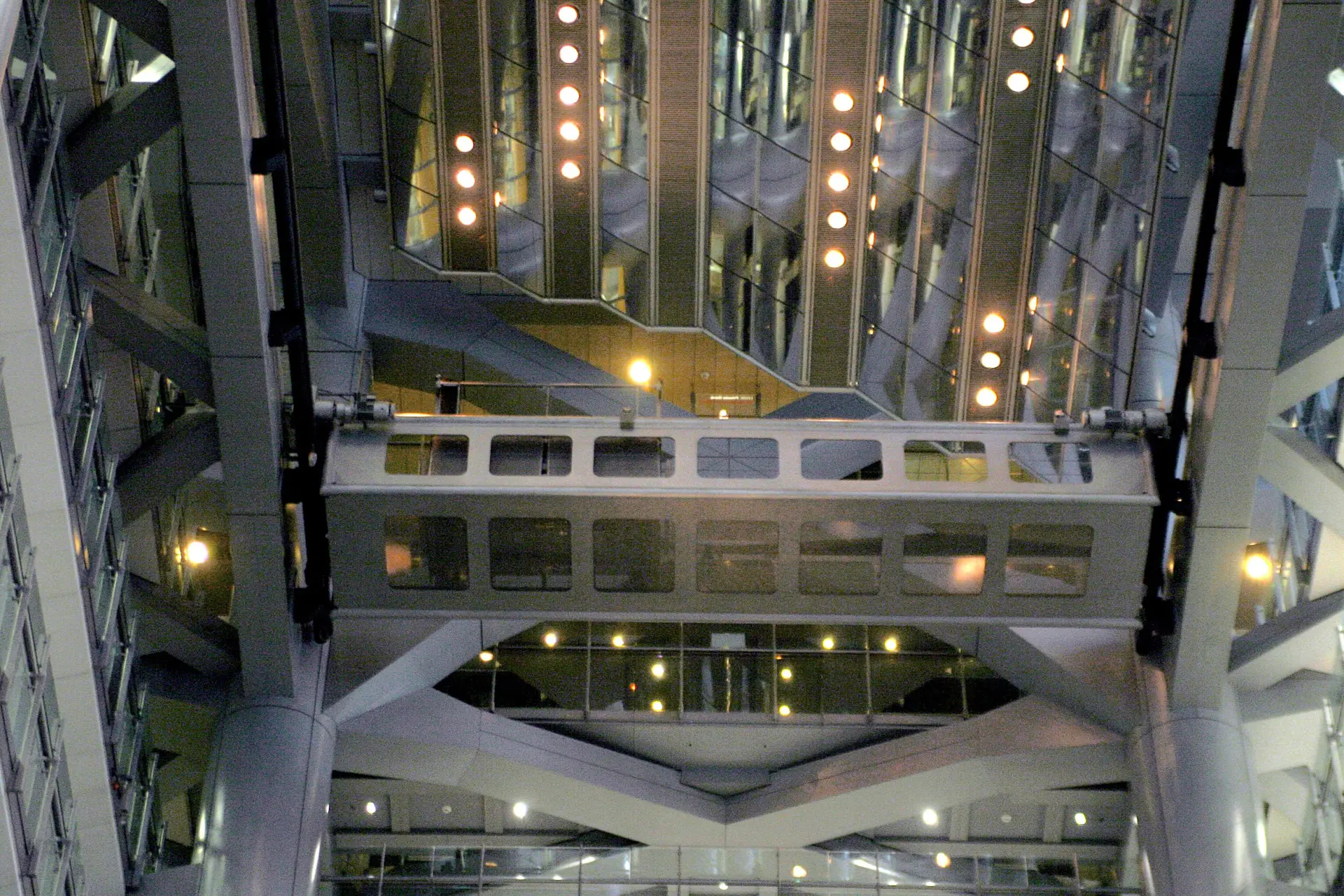 A modern glass atrium rises around intersecting steel walkways, with warm lights reflecting across the angular interior.