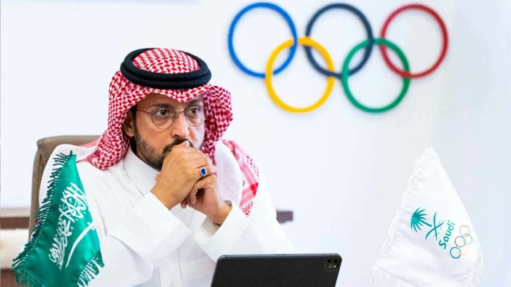 Prince Abdullah Bin Fahad, wearing a keffiyeh and glasses, sits at a desk with Saudi and Olympic flags, next to a large Olympic rings symbol.