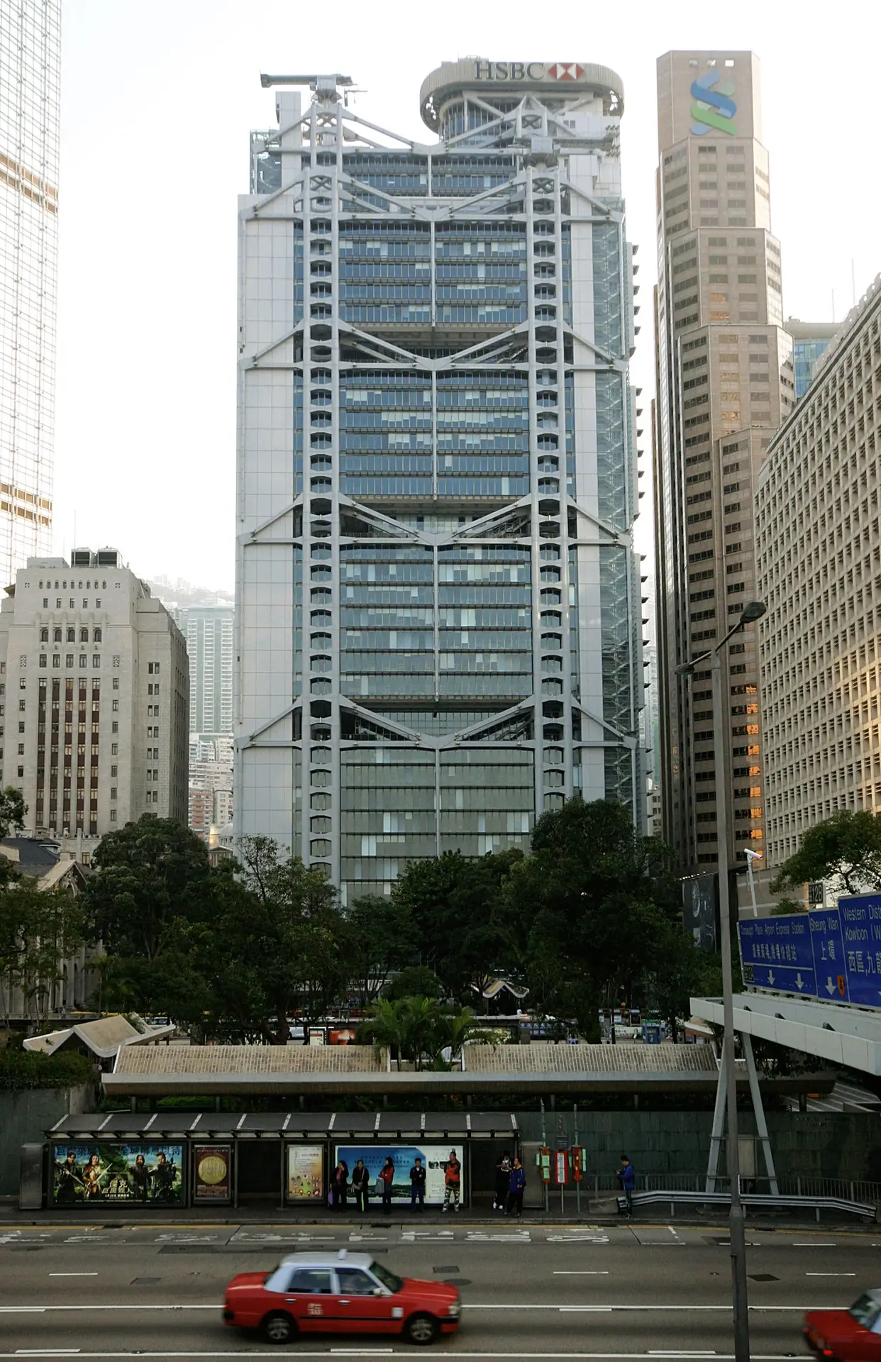 The HSBC tower rises above Hong Kong streets, with taxis passing in front and the Standard Chartered logo visible nearby.