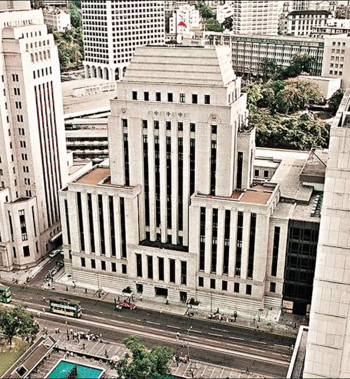 A cream-coloured civic building with tall vertical windows stands beside a busy road, framed by high-rise towers and trees.